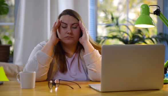Müde Frau im Homeoffice sitzt am Laptop, hat ihre Augen geschlossen und massiert sich die Schläfen.