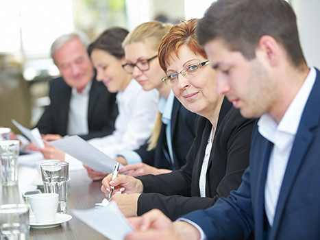 Frauen und Männer im Businessoutft sitzen in einer Reihe an einem Tisch bei einem Meeting, eine Frau schaut direkt in die Kamera.