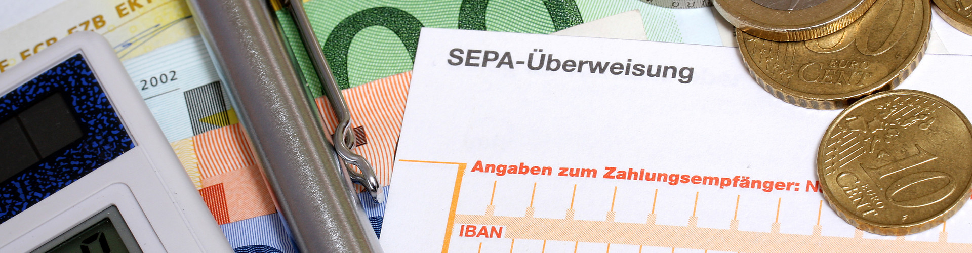 Auf mehreren Geldscheinen liegen Taschenrechner, Kugelschreiber, einige Euromünzen und ein Erlagschein. Zu erkennen ist die Aufschrift "SEPA-Überweisung". © RRF, stock.adobe.com
