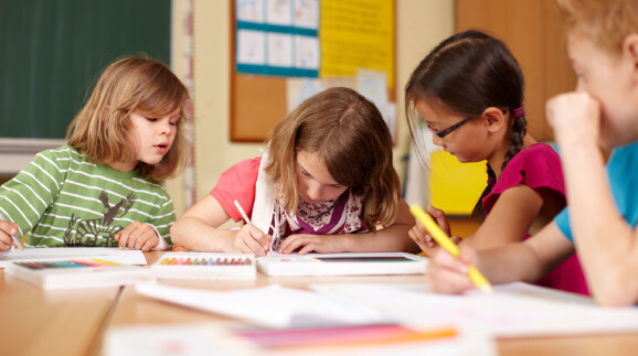 Vier Volksschulkinder sitzen nebeneinander an einer Schulbank und malen konzentriert