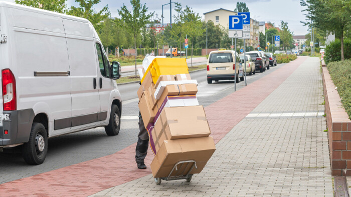 Ein Lieferbote transportiert auf einer Sackkarre einen hohen Stapel Kartons auf einem Gehweg neben einem wei&szlig;en Lieferwagen.