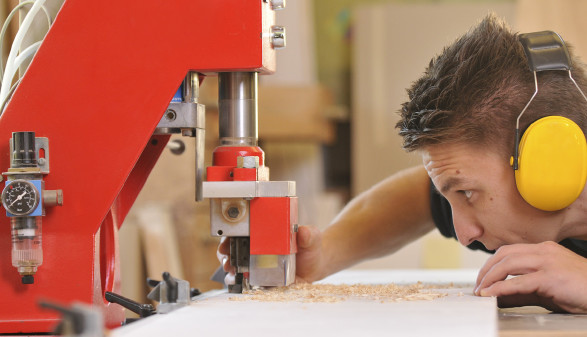 Ein männlicher Jugendlicher mit Gehörschutz steht an einer Werkbank und bearbeitet ein Holzbrett mit einer Fräsmaschine. © jörn buchheim, stock.adobe.com