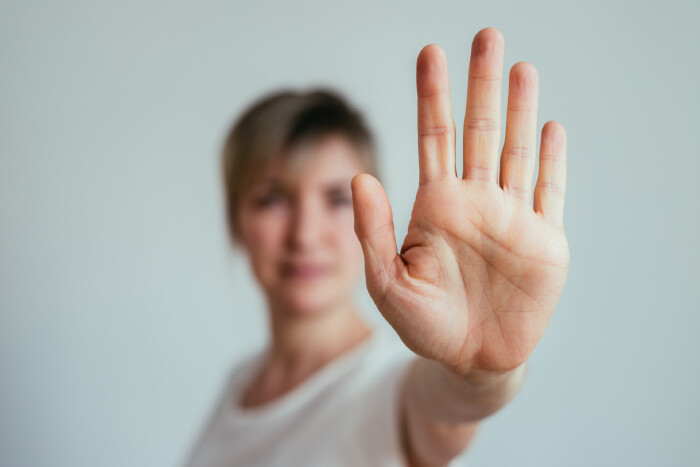 Frau mit wei&szlig;em T-Shirt unscharf vor wei&szlig;em Hintergrund abgebildet, sie hat ihren linken Arm in Richtung Kamera ausgestreckt und zeigt mit ihrer Hand eine scharf abgebildete Stopp-Geste