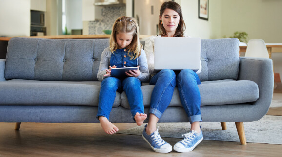 Mama mit Laptop und Kind mit Tablet sitzen auf dem Sofa.