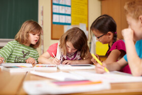 Vier Volksschulkinder sitzen nebeneinander an einer Schulbank und malen konzentriert
