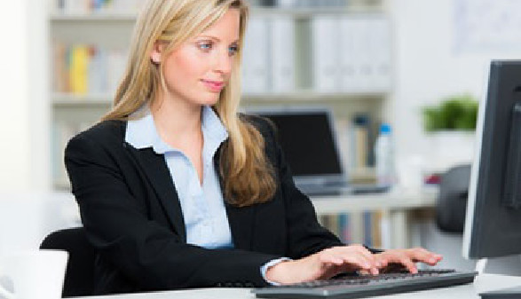 Eine Frau im Anzug sitzt am Schreibtisch und blickt auf den Computerbildschirm vor sich. Im Hintergrund sieht man ein Regal mit Ordnern.