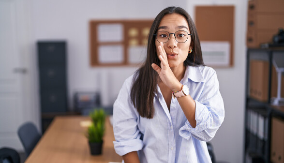 Junge Frau in einem Büro erzählt mit vorgehaltener Hand ein Geheimnis