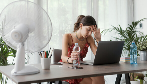 Junge Frau in leichtem Sommerkleid sitzt am Schreibtisch und wischt sich Schweiß von der Stirn. Vor ihr ein Ventilator und zwei Wasserflaschen.