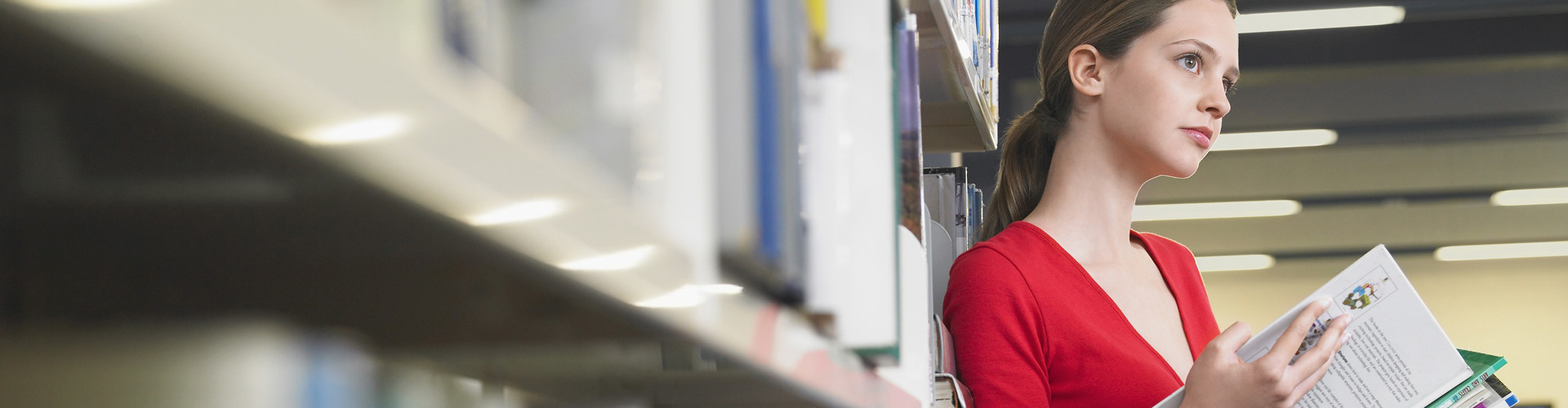 Junge Frau steht an ein Bücherregal gelehnt in der Bibliothek und hält dabei Lernunterlagen in der Hand. © biker3, stock.adobe.com