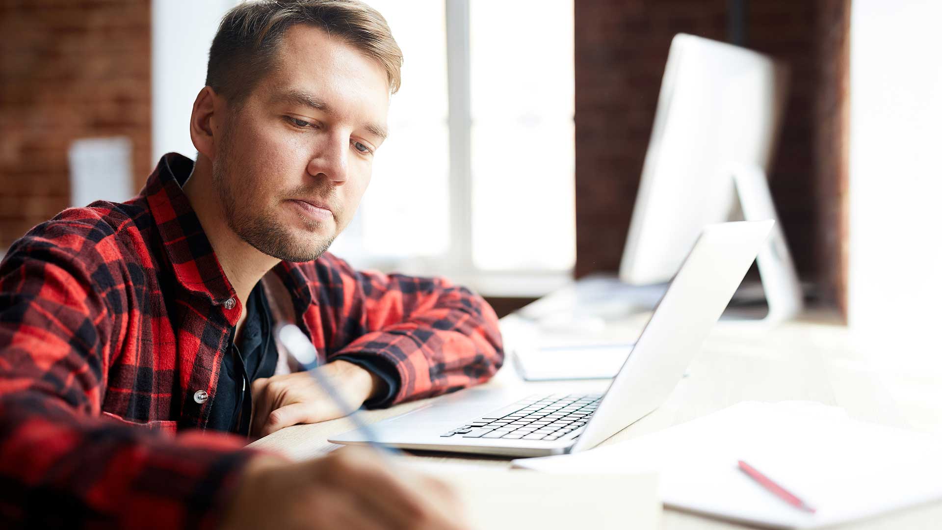 Junger Mann sitzt vor Laptop und macht sich Notizen auf seinem Block
